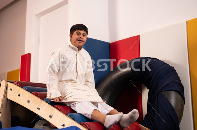 Spending a wonderful time at an amusement park, times of entertainment and fun, gestures of pleasure and happiness, enjoying summer vacation, a Saudi boy with Down syndrome at an amusement park, playing a fun slide game.