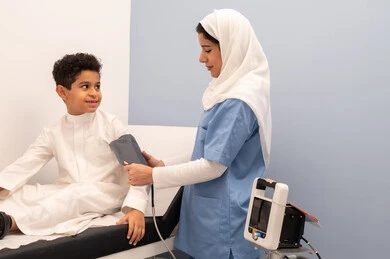 ممرضه سعودية تقوم بقياس ضغط الدم لولد عربي خليجي عماني يرتدي الزي العماني التقليدي يجلس على كرسي العيادة ، الشعور بالألم والوجع، متابعة دورية لفحص الضغط، تقديم الرعاية الصحية المنزلية، الفحص الطبي والتشخيص السريع، العمل في القطاع الصحي