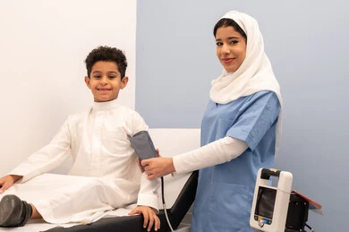 Working in the healthcare sector, feeling pain and discomfort, regular monitoring of blood pressure, providing home healthcare, medical examination and rapid diagnosis, a Saudi nurse measuring the blood pressure of an Arab Gulf Omani boy wearing traditional Omani attire sitting on a clinic chair.
