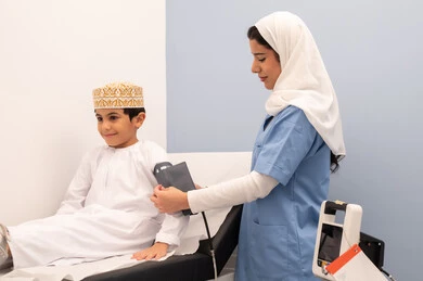 Medical examination and rapid diagnosis, feeling pain and discomfort, regular monitoring of blood pressure, providing home healthcare, working in the health sector, a Saudi nurse measuring the blood pressure of an Arab Gulf Omani boy wearing traditional Omani attire sitting on a clinic chair.