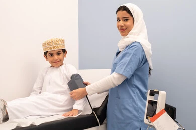 تقديم الرعاية الصحية المنزلية، الفحص الطبي والتشخيص السريع، الشعور بالألم والوجع، متابعة دورية لفحص الضغط،العمل في القطاع الصحي، ممرضه سعودية تقوم بقياس ضغط الدم لولد عربي خليجي عماني يرتدي الزي العماني التقليدي يجلس على كرسي العيادة