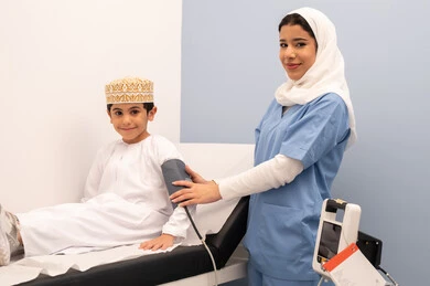 متابعة دورية لفحص الضغط، الشعور بالألم والوجع،تقديم الرعاية الصحية المنزلية، الفحص الطبي والتشخيص السريع، العمل في القطاع الصحي، ممرضه سعودية تقوم بقياس ضغط الدم لولد عربي خليجي عماني يرتدي الزي العماني التقليدي يجلس على كرسي العيادة