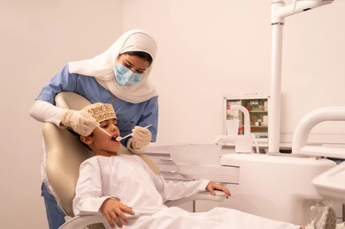 طبيبة ترتدي القفازات تقوم بفحص أسنان المريض، ولد عربي خليجي عماني يرتدي الزي العماني التقليدي  يجلس على كرسي العلاج في العيادة السنية،مفهوم الطب والرعاية الصحية، استخدام المعدات اللازمة لجراحة الاسنان ،  تقديم الخدمات الطبية للمرضى ، إجراء الفحوصات الدورية للعناية بالصحة الفموية