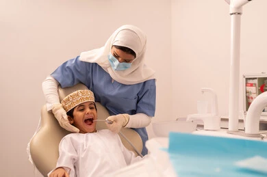 Providing medical services to patients, an Omani Gulf Arab boy wearing traditional Omani attire sitting on the treatment chair in the dental clinic, the concept of medicine and healthcare, using the necessary equipment for dental surgery, a female doctor wearing gloves examining the patient's teeth, conducting regular check-ups for oral health care.