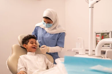 إجراء الفحوصات الدورية للعناية بالصحة الفموية، مفهوم الطب والرعاية الصحية، استخدام المعدات اللازمة لجراحة الاسنان ، طبيبة ترتدي القفازات تقوم بفحص أسنان المريض، ولد عربي خليجي عماني يرتدي الزي العماني التقليدي يجلس على كرسي العلاج في العيادة السنية، تقديم الخدمات الطبية للمرضى