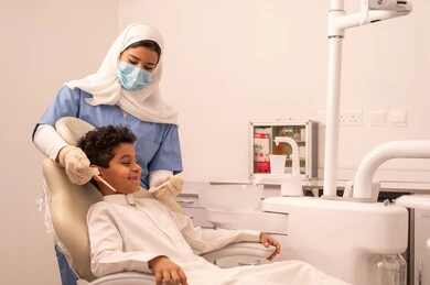The use of necessary equipment for dental surgery, the concept of medicine and healthcare, a female doctor wearing gloves examining a patient's teeth, an Arab Gulf Omani boy wearing traditional Omani attire sitting on the treatment chair in the dental clinic, providing medical services to patients, conducting regular check-ups for oral health care.