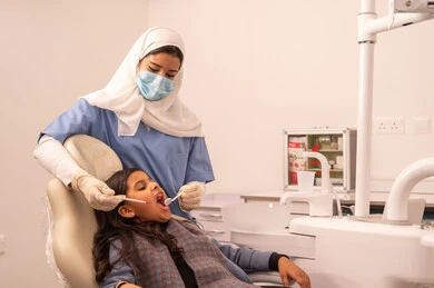 Conducting regular check-ups for oral health care, using the necessary equipment for dental surgery, the concept of medicine and healthcare, a doctor wearing gloves examining a patient's teeth, an Arab Gulf Omani girl wearing casual attire sitting on the treatment chair in the dental clinic, providing medical services to patients.