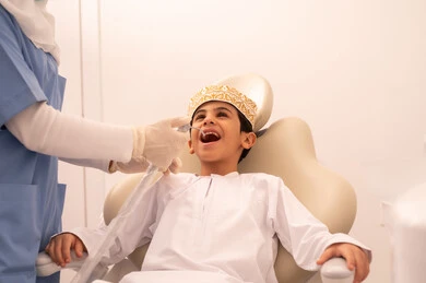 طبيبة ترتدي القفازات تقوم بفحص أسنان المريض، مفهوم الطب والرعاية الصحية، استخدام المعدات اللازمة لجراحة الاسنان ، ولد عربي خليجي عماني يرتدي الزي العماني التقليدي  يجلس على كرسي العلاج في العيادة السنية، تقديم الخدمات الطبية للمرضى ، إجراء الفحوصات الدورية للعناية بالصحة الفموية