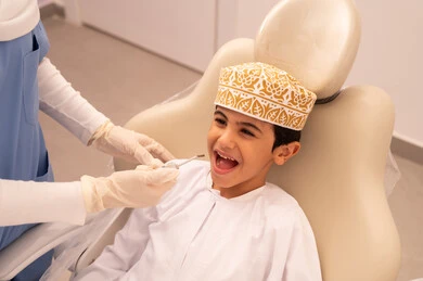 The concept of medicine and healthcare, the use of necessary equipment for dental surgery, a female doctor wearing gloves examining a patient's teeth, an Arab Gulf Omani boy wearing traditional Omani attire sitting on the treatment chair in the dental clinic, providing medical services to patients, conducting regular check-ups for oral health care.