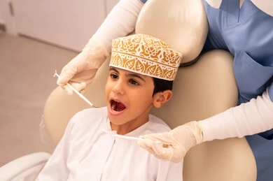 The use of necessary equipment for dental surgery, the concept of medicine and healthcare, a female doctor wearing gloves examining a patient's teeth, an Arab Gulf Omani boy wearing traditional Omani attire sitting on the treatment chair in the dental clinic, providing medical services to patients, conducting regular check-ups for oral health care.