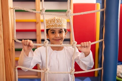 تنمية المهارات الحركية للأطفال، ولد عربي خليجي عماني يرتدي الدشداشة والكمه يستمتع باللعب في مدينة الألعاب ، يتسلق على الألعاب الخشبية المجهزة بالحبال والعوارض الملونة، قضاء أوقات ممتعة مليئة بالنشاط والحركة، مفهوم المرح والترفيه