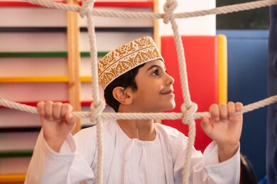 ولد عربي خليجي عماني يرتدي الدشداشة والكمه يستمتع باللعب في مدينة الألعاب ، يتسلق على الألعاب الخشبية المجهزة بالحبال والعوارض الملونة، قضاء أوقات ممتعة مليئة بالنشاط والحركة، تنمية المهارات الحركية للأطفال، مفهوم المرح والترفيه