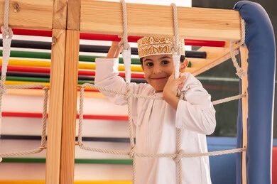 مفهوم المرح والترفيه ،تنمية المهارات الحركية للأطفال، ولد عربي خليجي عماني يرتدي الدشداشة والكمه يستمتع باللعب في مدينة الألعاب ، يتسلق على الألعاب الخشبية المجهزة بالحبال والعوارض الملونة، قضاء أوقات ممتعة مليئة بالنشاط والحركة