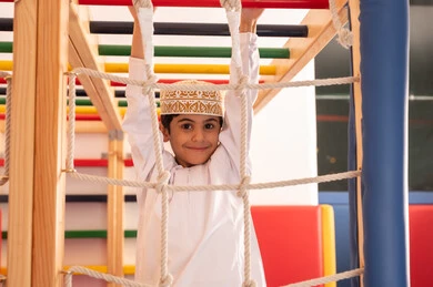 قضاء أوقات ممتعة مليئة بالنشاط والحركة، تنمية المهارات الحركية للأطفال، ولد عربي خليجي عماني يرتدي الدشداشة والكمه يستمتع باللعب في مدينة الألعاب ، يتسلق على الألعاب الخشبية المجهزة بالحبال والعوارض الملونة ،مفهوم المرح والترفيه