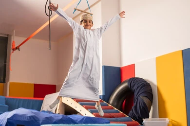 Enjoyable summer atmosphere, amusement city in the Sultanate of Oman, the concept of fun and play, jumping high with gestures of happiness and joy, an Arab Gulf Omani boy wearing a dishdasha and kumma enjoying playing in front of the slide, enjoying recreational games, games of entertainment and fun.