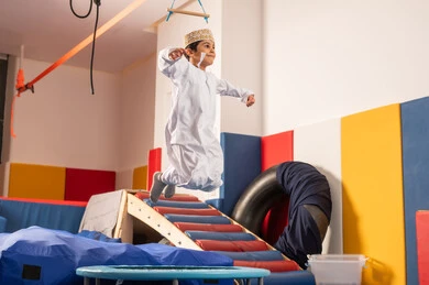 القفز عالياً بايماءات السعادة والسرور، مفهوم اللهو واللعب،اجواء صيفية ممتعة، مدينه الالعاب في سلطنة عمان، ولد عربي خليجي عماني يرتدي الدشداشه والكمه مستمتع باللعب امام الزحليقة ، الاستمتاع بالالعاب الترفيهية، العاب التسلية والمتعه