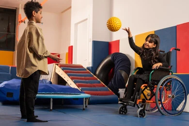 An Omani girl sitting in a wheelchair playing with a ball in an amusement park accompanied by a Saudi boy, engaging in many fun activities for people with determination, taking care of children with special needs, kindergartens, and nurseries.