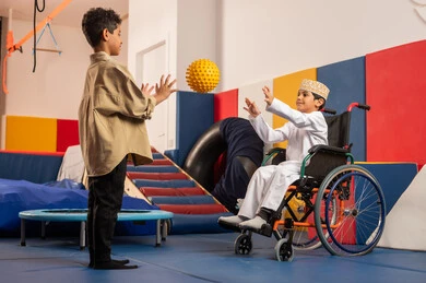 Two Omani boys are playing ball in the amusement park, one of them is sitting in a wheelchair, performing many fun activities for people of determination, taking care of children with special needs, kindergartens, and nurseries.
