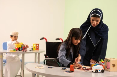 An Omani boy is playing with plastic toys, engaging in many fun activities for people with determination. An Arab Omani teacher for kindergarten specializes in caring for children with special needs. In the kindergartens and nurseries, an Arab Gulf Omani girl wearing casual attire is performing skills and activities alongside her teacher.