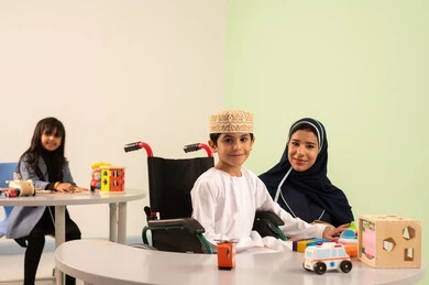 A Gulf Arab Omani boy wearing a dishdasha and kumma is performing skills and activities alongside his teacher, engaging in many fun activities for individuals with special needs. An Omani Arab kindergarten teacher specializes in caring for children with special needs, kindergartens, and nurseries. An Omani girl is playing with plastic toys.