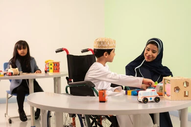 أداء الكثير من الأنشطة الممتعة لذوي الهمم، معلمة عربية عمانيه لرياض أطفال تختص بالاعتناء بطفل ذوي الاحتياجات الخاصة، رياض الأطفال والحاضنات، ولد عربي خليجي عماني يرتدي الدشداشه والكمه يقوم بالمهارات والانشطه برفقه معلمته ، بنت عمانية تلعب بالالعاب البلاستيكيه