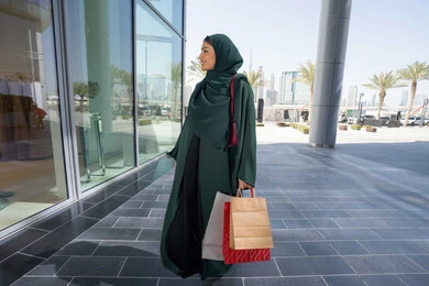 Excited to take advantage of discounts on products, a veiled Emirati Gulf Arab woman wearing an abaya holding paper bags, taking advantage of seasonal offers and discounts and White Friday, shopping in malls, gestures of happiness and pleasure