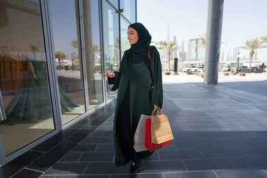 السعادة والتأثير الإيجابي على السيدات في زيارة المولات التجارية ، إمرأة عربية خليجية اماراتية محجبة ترتدي العباءه تحمل بيدها أكياس ورقية, استغلال العروض والتخفيضات الموسمية والجمعة البيضاء، التسوق من المولات التجارية، ايماءات السعاده و السرور