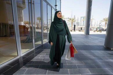 عروض وتخفيضات موسمية في الأسواق الاماراتية، التبضع وأخذ المستلزمات الضرورية، إمرأة عربية خليجية اماراتية محجبة ترتدي العباءه تحمل بيدها أكياس ورقية, استغلال العروض والتخفيضات الموسمية والجمعة البيضاء، ايماءات السعاده و السرور