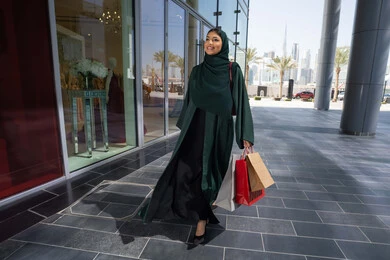 شراء الهدايا والمستلزمات، الترويج لماركات عالمية فاخرة ، إمرأة عربية خليجية اماراتية محجبة ترتدي العباءه تحمل بيدها أكياس ورقية, استغلال العروض والتخفيضات الموسمية والجمعة البيضاء، التسوق من المولات التجارية، ايماءات السعاده و السرور