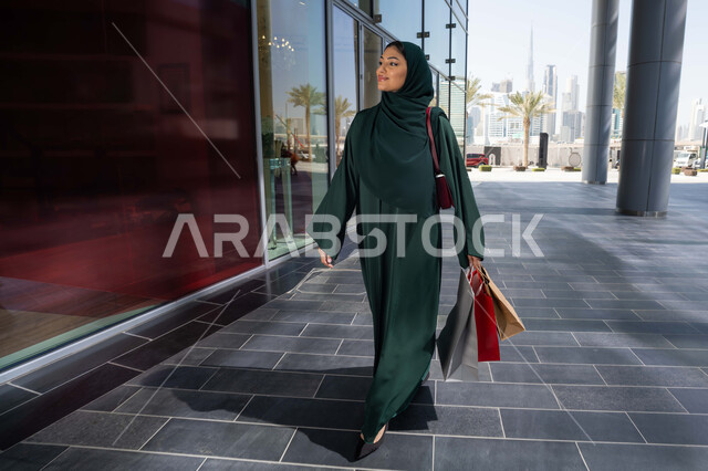 Choosing things and supplies from shopping malls, a veiled Emirati Gulf Arab woman wearing an abaya holding paper bags, taking advantage of seasonal offers and discounts and White Friday, shopping from shopping malls, gestures of happiness and pleasure