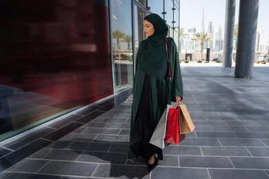 الاستمتاع بقضاء وقت ممتع في التسوق والشراء، إمرأة عربية خليجية اماراتية محجبة ترتدي العباءه تحمل بيدها أكياس ورقية, استغلال العروض والتخفيضات الموسمية والجمعة البيضاء، التسوق من المولات التجارية، ايماءات السعاده و السرور