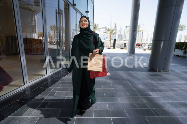 ايماءات السعاده و السرور، متعة الشراء من الماركات العالمية، إمرأة عربية خليجية اماراتية محجبة ترتدي العباءه تحمل بيدها أكياس ورقية, استغلال العروض والتخفيضات الموسمية والجمعة البيضاء، التسوق من المولات التجارية