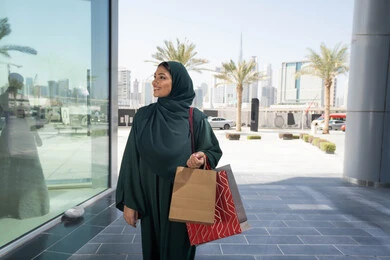 التبضع وأخذ المستلزمات الضرورية،  إمرأة عربية خليجية اماراتية محجبة ترتدي العباءه تحمل بيدها أكياس ورقية, استغلال العروض والتخفيضات الموسمية والجمعة البيضاء، التسوق من المولات التجارية، ايماءات السعاده و السرور