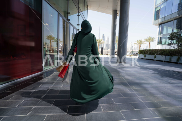 خلفية خارج المول التجاري، الاستمتاع بالشراء والتسوق ،صورة من الخلف لامراة عربية اماراتية خليجية ترتدي العباءة تحمل بيديها حقائب الشراء، ايماءات الوجه تدل على السعادة ، قضاء وقت ممتع في شراء المستلزمات ، الاستفادة من العروض والخصومات