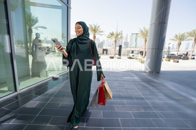 اقتناء بعض المستلزمات والهدايا، استخدام الأجهزة والتقنيات الحديثة والمتطورة, تصفح تطبيقات الجوال لشراء المنتجات، سيدة عربية خليجية اماراتية ترتدي الحجاب والعباءة تمسك بيدها الهاتف المحمول وأكياس التسوق