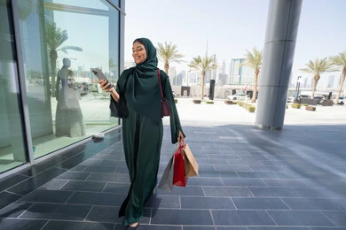 اقتناء بعض المستلزمات والهدايا، استخدام الأجهزة والتقنيات الحديثة والمتطورة, تصفح تطبيقات الجوال لشراء المنتجات، سيدة عربية خليجية اماراتية ترتدي الحجاب والعباءة تمسك بيدها الهاتف المحمول وأكياس التسوق