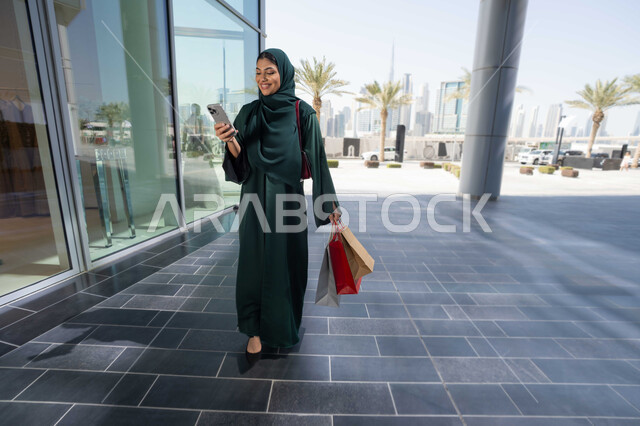 التبضع والتسوق من الماركات العالميه ، الجمعه البيضاء، استخدام الأجهزة والتقنيات الحديثة والمتطورة, تصفح تطبيقات الجوال لشراء المنتجات، سيدة عربية خليجية اماراتية ترتدي الحجاب والعباءة تمسك بيدها الهاتف المحمول وأكياس التسوق