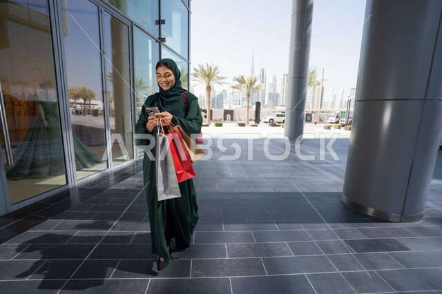 شراء الاحتياجات والملابس من المولات التجاريه ،استغلال العروض الكبرى، استخدام الأجهزة والتقنيات الحديثة والمتطورة, تصفح تطبيقات الجوال لشراء المنتجات، سيدة عربية خليجية اماراتية ترتدي الحجاب والعباءة تمسك بيدها الهاتف المحمول وأكياس التسوق