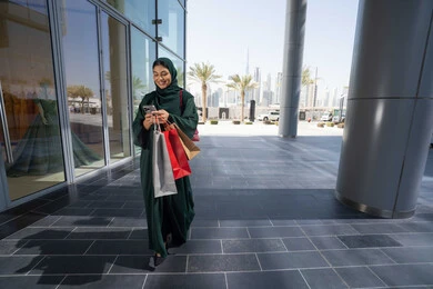 شراء الاحتياجات والملابس من المولات التجاريه ،استغلال العروض الكبرى، استخدام الأجهزة والتقنيات الحديثة والمتطورة, تصفح تطبيقات الجوال لشراء المنتجات، سيدة عربية خليجية اماراتية ترتدي الحجاب والعباءة تمسك بيدها الهاتف المحمول وأكياس التسوق