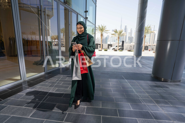 استغلال العروض الكبرى, شراء الاحتياجات والملابس من المولات التجاريه، استخدام الأجهزة والتقنيات الحديثة والمتطورة, تصفح تطبيقات الجوال لشراء المنتجات، سيدة عربية خليجية اماراتية ترتدي الحجاب والعباءة تمسك بيدها الهاتف المحمول وأكياس التسوق