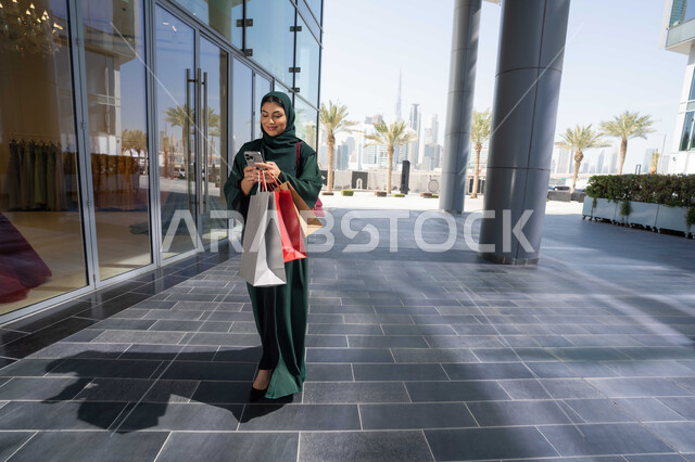 التسوق وشراء الحاجيات والمستلزمات التموينيه من المراكز التجارية، استخدام الأجهزة والتقنيات الحديثة والمتطورة, تصفح تطبيقات الجوال لشراء المنتجات، سيدة عربية خليجية اماراتية ترتدي الحجاب والعباءة تمسك بيدها الهاتف المحمول وأكياس التسوق