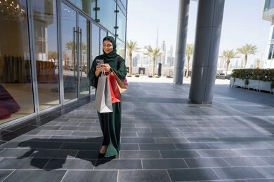 Shopping and purchasing necessities and supplies from malls, using modern and advanced devices and technologies, browsing mobile applications to purchase products, an Emirati Gulf Arab woman wearing a hijab and abaya holding a mobile phone and shopping bags in her hand