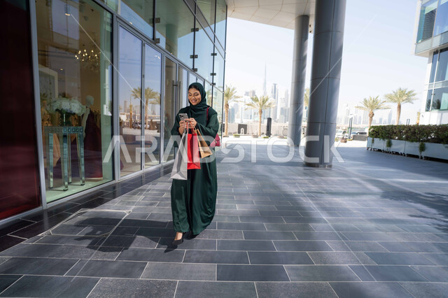 شراء الأغراض الجديدة من المول، استخدام الأجهزة والتقنيات الحديثة والمتطورة، سيدة عربية خليجية اماراتية ترتدي الحجاب والعباءة تمسك بيدها الهاتف المحمول وأكياس التسوق