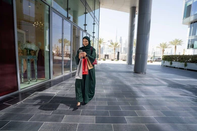 شراء الأغراض الجديدة من المول، استخدام الأجهزة والتقنيات الحديثة والمتطورة، سيدة عربية خليجية اماراتية ترتدي الحجاب والعباءة تمسك بيدها الهاتف المحمول وأكياس التسوق