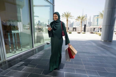 متعة الشراء من الماركات العالمية، استخدام الأجهزة والتقنيات الحديثة والمتطورة، سيدة عربية خليجية اماراتية ترتدي الحجاب والعباءة تمسك بيدها الهاتف المحمول وأكياس التسوق