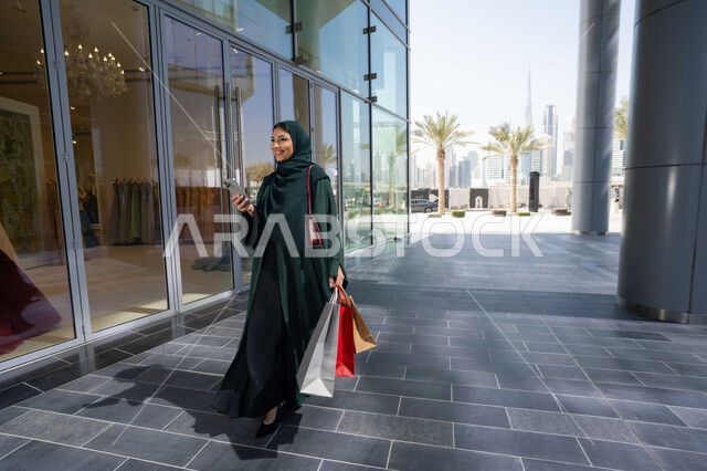 شراء المستلزمات والتبضع من المولات التجارية، استخدام الأجهزة والتقنيات الحديثة والمتطورة، سيدة عربية خليجية اماراتية ترتدي الحجاب والعباءة تمسك بيدها الهاتف المحمول وأكياس التسوق