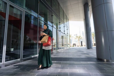 تصفح تطبيقات الجوال لشراء المنتجات، استخدام الأجهزة والتقنيات الحديثة والمتطورة، سيدة عربية خليجية اماراتية ترتدي الحجاب والعباءة تمسك بيدها الهاتف المحمول وأكياس التسوق