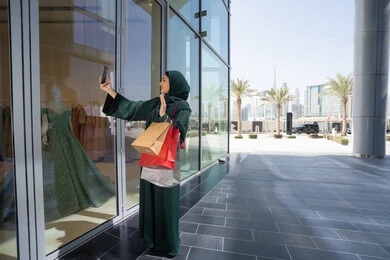 متعة التسوق في الامارات، استخدام التكنولوجيا في الحياة اليومية، إمرأة سعودية تقوم بالتسوق وشراء المستلزمات والهدايا عبر الانترنت،سيدة عربية خليجية اماراتية ترتدي الحجاب والعباءة تمسك بيدها الهاتف المحمول وأكياس التسوق، التقاط صور السيلفي التذكارية