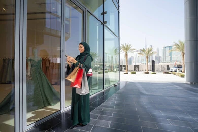 استخدام الأجهزة والتقنيات الحديثة والمتطورة, تصفح تطبيقات الجوال لشراء المنتجات، سيدة عربية خليجية اماراتية ترتدي الحجاب والعباءة تمسك بيدها الهاتف المحمول وأكياس التسوق