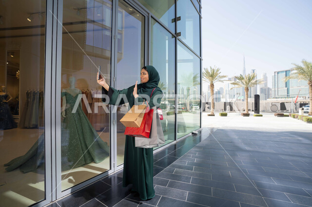 Taking selfies with a mobile phone, taking advantage of seasonal offers and discounts, using technology in daily life, a Saudi woman shopping and buying supplies and gifts online, an Emirati Gulf Arab woman wearing a hijab and abaya holding a mobile phone and shopping bags